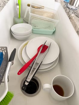 Castle Rock, Colorado, USA-January 18, 2024-A white kitchen sink filled with dirty dishes including plates bowls a coffee mug and utensils such as tongs and knives A dish rack with soapy water is placed on one side while a sponge and dish scrubber rest nearby The sink has a modern design with a garbage disposal drain