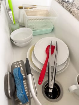 Castle Rock, Colorado, USA-January 18, 2024-A white kitchen sink filled with dirty dishes including plates bowls a coffee mug and utensils such as tongs and knives A dish rack with soapy water is placed on one side while a sponge and dish scrubber rest nearby The sink has a modern design with a garbage disposal drain