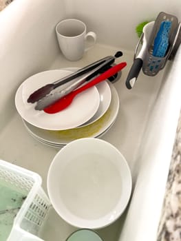 Castle Rock, Colorado, USA-January 18, 2024-A white kitchen sink filled with dirty dishes including plates bowls a coffee mug and utensils such as tongs and knives A dish rack with soapy water is placed on one side while a sponge and dish scrubber rest nearby The sink has a modern design with a garbage disposal drain