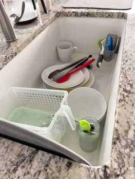 Castle Rock, Colorado, USA-January 18, 2024-A white kitchen sink filled with dirty dishes including plates bowls a coffee mug and utensils such as tongs and knives A dish rack with soapy water is placed on one side while a sponge and dish scrubber rest nearby The sink has a modern design with a garbage disposal drain