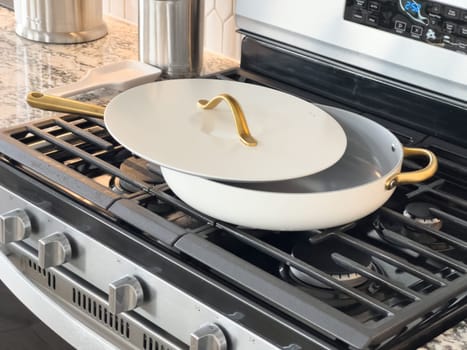 Castle Rock, Colorado, USA-January 18, 2025-A white cooking pan with gold handles and a matching lid sits on a gas stove The pan has a sleek modern design with a nonstick surface The stainless steel stove has multiple burners and a digital display The kitchen counter features a marble design with various cooking utensils in the background