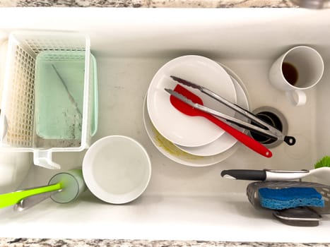 Castle Rock, Colorado, USA-January 18, 2024-A white kitchen sink filled with dirty dishes including plates bowls a coffee mug and utensils such as tongs and knives A dish rack with soapy water is placed on one side while a sponge and dish scrubber rest nearby The sink has a modern design with a garbage disposal drain
