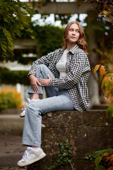 A young woman with wavy brown hair and blue eyes poses in a serene autumn park, sitting on a rustic stone wall surrounded by lush green and orange foliage. She wears a black and white checkered shirt over a white top, paired with light blue jeans and white sneakers. Her confident and relaxed expression, combined with soft natural lighting, creates a harmonious and stylish outdoor portrait. This image is ideal for autumn fashion promotions, lifestyle blogs, seasonal advertising, and social media campaigns.
