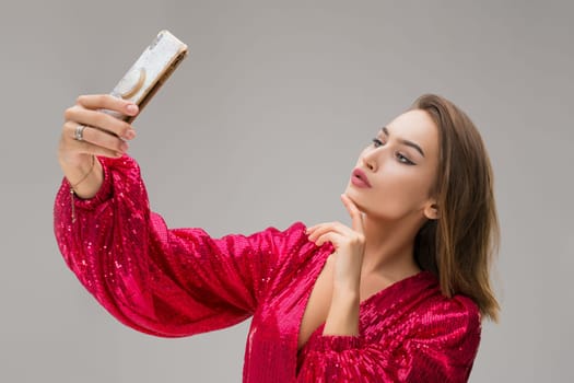 Cheerful young female in festive red dress doing selfie on modern smartphone with bright shiny design while standing against gray background