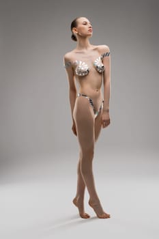 Full body slim woman with bra and panties made of metallic tape looking away and tiptoeing against gray background