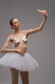 Attractive young slim female in ballet tutu skirt and with sticky tapes on breast taking selfie on smartphone against gray background