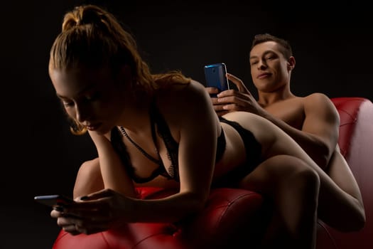 Young seductive woman in sexy lingerie and handsome brutal boyfriend browsing mobile phones while relaxing on red leather chair in dark studio