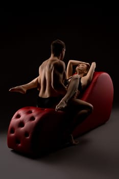 Sexy muscular man sitting before red woman in black lingerie leaning on red armchair during foreplay in studio
