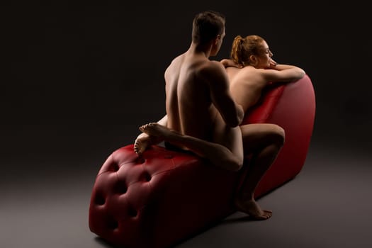 Sexy muscular man sitting before red woman in black lingerie leaning on red armchair during foreplay in studio
