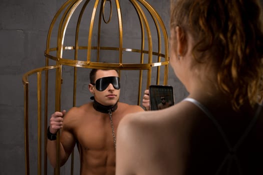 Unrecognizable woman with smartphone taking picture of male lover in black eyes cover with chain and handcuffs fastened to metal cage during sexual BDSM game