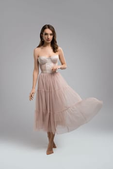 Full body of charming tender young female model wearing white corset and tulle skirt looking at camera while standing in light studio