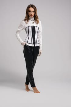 Full body slim young woman in elegant outfit with corset holding hand in pocket and looking at camera while tiptoeing against gray background