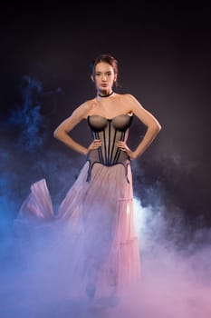Charming female wearing corset and maxi dress standing in studio with smoke on dark background and looking at camera