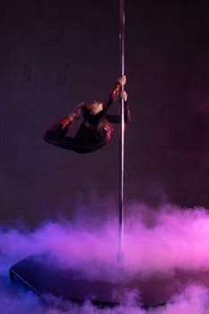 Full body side view of flexible teen female dancer doing aerial trick on pylon during performance on stage with smoke and neon illumination