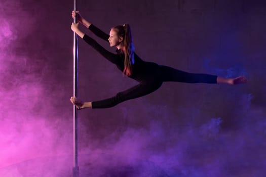 Full body side view of young flexible teen female dancer in black hanging on pole and doing splits during performance under neon light and smoke
