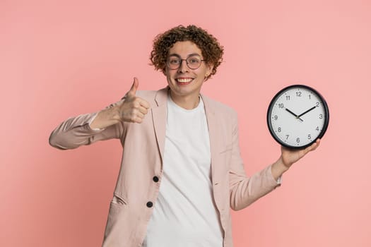 It is your time, hurry up. Caucasian man showing time on wall office clock, ok thumb up, approve, pointing finger at camera, advertisement, deadline. Curly haired young guy isolated on pink background