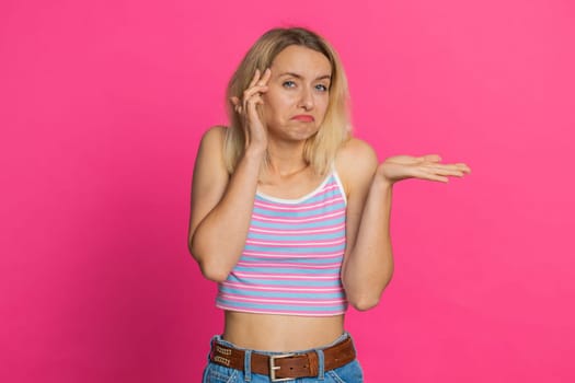 I don't know what to say. Confused Caucasian young woman feeling embarrassed about ambiguous question, having doubts, no answer, idea, being clueless uncertain. Blonde girl isolated on pink background