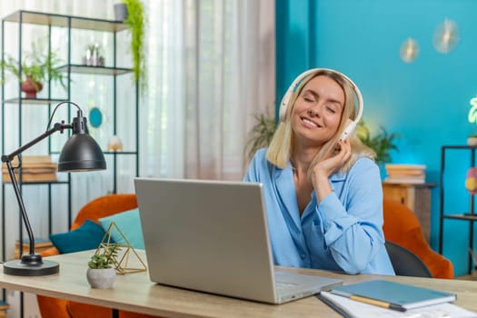 Happy overjoyed businesswoman sitting at home office desk listening to favorite energetic disco music through wireless headphones. Happy freelancer girl relaxing, taking a break with laptop on table.