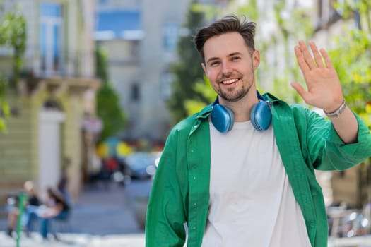 Cheerful young man tourist smiling friendly at camera, waving hands gesturing invitation hello, hi, greeting, goodbye, welcoming with hospitable expression outdoors. Guy on urban city town street.