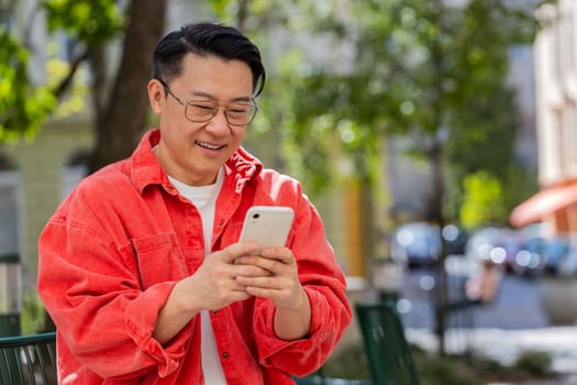 Asian middle-aged man using smartphone typing text messages in social media application online, surfing internet, relaxing, taking a break outdoors. Happy Chinese guy sitting on urban city street.