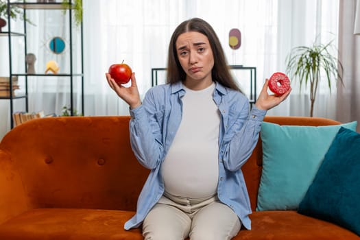 Choosing right nutrition. Confused young pregnant woman comparing sweet donut and ripe apple and shrugging shoulders in uncertainty. Future mother girl hesitates choosing between dessert and fruit