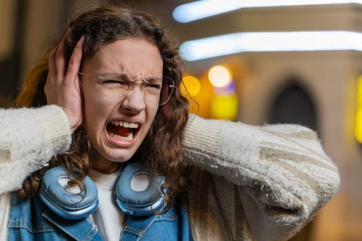 Displeased angry woman frowns face feels disgust suffering from loud sound covers ears with hands palms refusing to listen gossip bad information don't hear. Screaming. Girl stand on street at night.