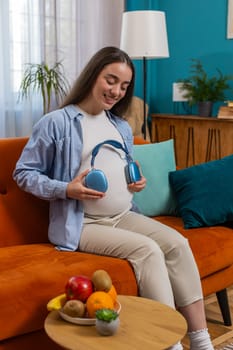 Happy pregnant woman girl sitting on sofa putting wireless headphones on her tummy with child baby while listening to favorite music at home. Technology, future mother and domestic activity concept.
