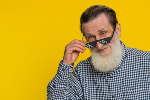 Portrait of stylish senior old man wearing black sunglasses charming smile, celebrating success victory win good positive news. Happy elderly grandfather pensioner on yellow background. Wink blink eye