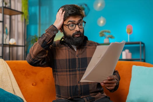 Frustrated upset amazed Indian business man works at home sofa looking at bills paperwork reports documents gets angry, frustrated, stress bankruptcy financial problem. Stresses freelancer at home