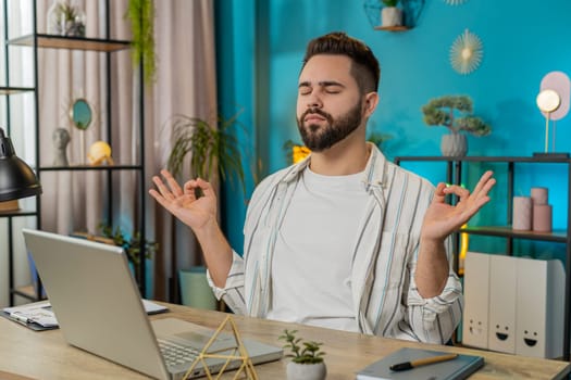 Peaceful relaxed male worker businessman sitting at workplace with folded in mudra gesture hands, reducing stress during workday, meditating or doing yoga breathing exercises in home office. Lifestyle