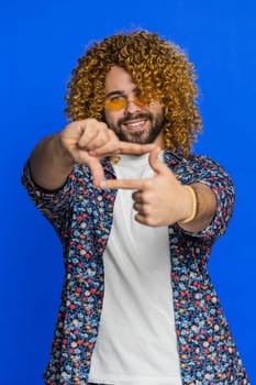 Man photographer gesturing picture frame with hands, looks through fingers and focusing on interesting moment, imitating zoom and cropping nice image. Young guy isolated on blue background. Vertical