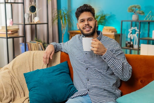 Smiling Indian Hindu man drinking a cup of warm coffee or herbal tea sitting at home living room couch in morning. Hispanic guy enjoying comfortable relaxing alone. Relaxing, taking a break. Lifestyle