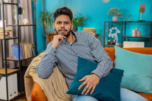 Portrait of happy calm Hindu man at home couch smiling friendly, glad expression looking away dreaming resting, relaxation feel satisfied good news, celebrate win. Hispanic young guy in living room