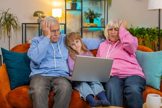 Upset grandfather, grandmother and granddaughter playing game on laptop and losing at home. Sad Caucasian girl with grandparents received bad news, fortune loss, fail education study test on couch