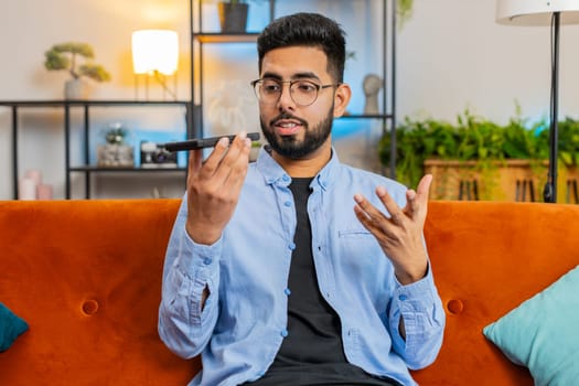 Cheerful Indian man enjoying mobile loudspeaker talking gossip, rumors, good news sitting on sofa. Happy excited Arabian guy making phone call conversation with friends at modern home room apartment.