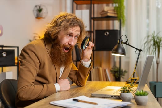 Surprised investigator researcher businessman working at home office holding magnifying glass looking at laptop screen with wow expression, searching, analyzing. Excited freelancer at home office.