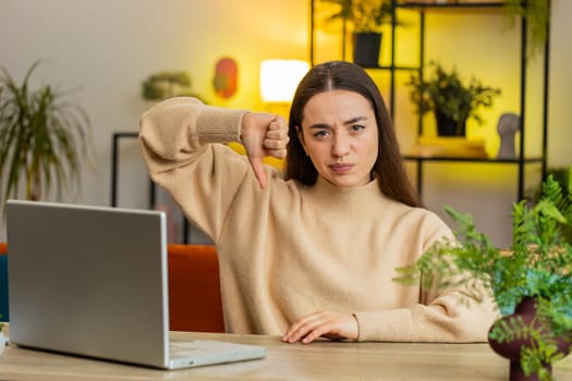Dislike. Upset unhappy Caucasian woman showing thumb down sign gesture expressing discontent disapproval dissatisfied bad work at home office indoors. Displeased pretty young girl in room at table