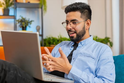 Full length of smiling Indian man typing on laptop using apps lying on comfortable sofa. Happy young Arabian guy working on computer in living room at home. E-learning, browsing internet on netbook