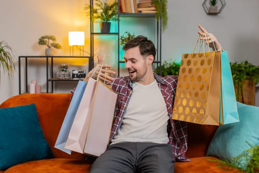 Young happy man shopaholic consumer came back home after shopping sale with bags. Discount sellout on mall Caucasian guy satisfied received parcels purchase from online order at home apartment on sofa