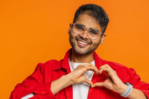 Man in love. Smiling handsome Indian man makes heart gesture demonstrates love sign expresses good positive feelings and sympathy. Handsome Arabian Hindu young guy isolated on orange wall background