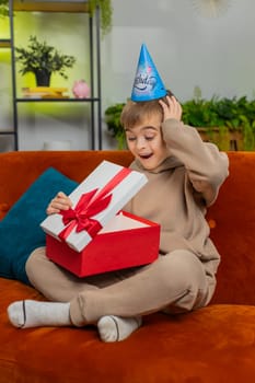 Happy preteen child boy wears festive birthday cap hat hold gift box with ribbon congratulating. Teenager kid celebrating party event opening delivery greeting present at home room on couch. Vertical