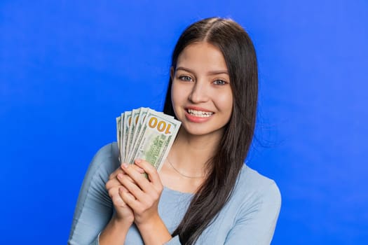 Rich pleased happy Caucasian smiling woman waving money dollar cash banknotes bills like a fan, success business career, lottery winner, big income wealth. Teenager girl isolated on blue background
