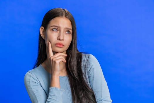 Thoughtful clever Caucasian young woman rubbing her chin and looking aside with pensive expression, pondering a solution strategy, doubting question. Girl isolated on studio blue background indoors