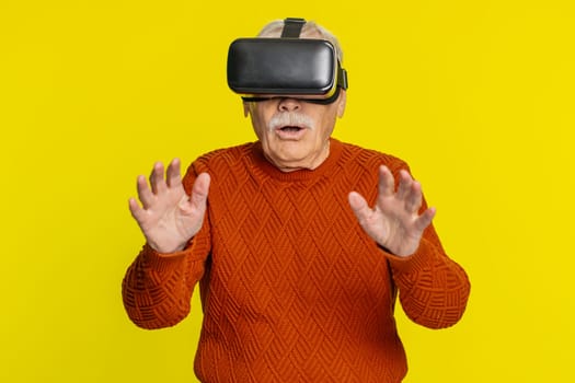 Excited happy senior old man using headset helmet app to play simulation game. Watching virtual reality 3D 360 video. Elderly grandfather in VR goggles isolated on yellow background. Future technology