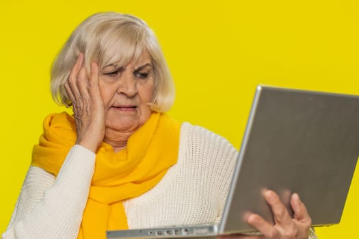 Displeased senior woman use laptop notebook typing browsing working loses becoming surprised by lottery bad results news fortune lost game fail computer virus. Mature grandmother on yellow background