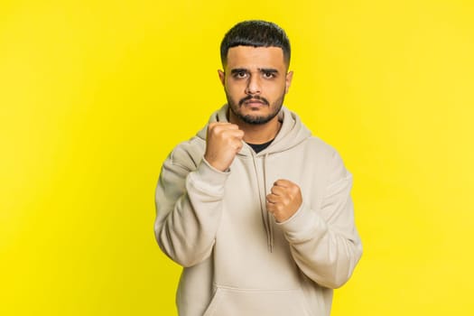 Aggressive angry Indian young man trying to fight at camera, shaking fist, boxing with expression, punishment, threaten, bullying, abuse, mad fury. Arabian guy isolated on yellow studio background