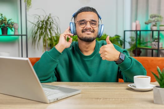 Happy relaxed Indian man in wireless headphones dancing at home office table workplace choosing listening favorite energetic disco music entertaining showing thumbs up. People weekend activities