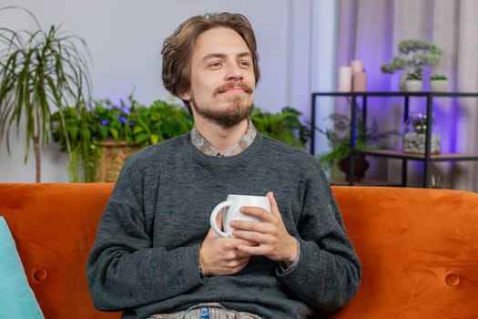 Attractive smiling bearded man drinking a cup of warm coffee or herbal tea sitting at home apartment living room couch in the morning. Caucasian young adult guy enjoying comfortable relaxing alone