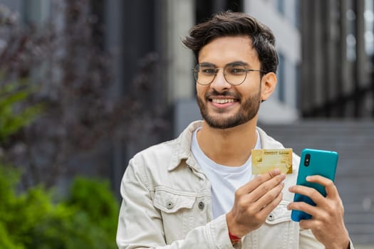 Happy Indian man using credit bank card smartphone while transferring money, purchases online shopping order food delivery, booking outdoors. Arabian Hindu guy in downtown city street. Business people