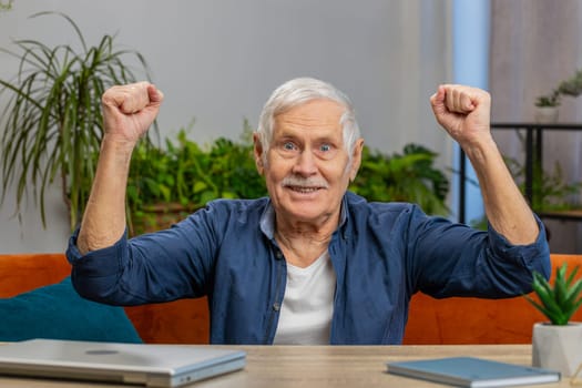 Happy senior man celebrating success, winning, goal achievement good news, lottery luck, victory, watching TV championship at modern home. Elderly mature grandfather in living room sitting on table